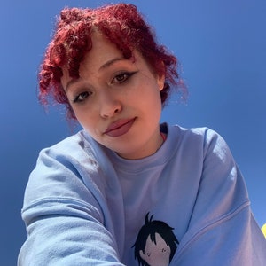 May include: A person with red curly hair is wearing a light blue sweatshirt with a black and white cartoon character embroidered on the front. The character has black hair and a simple face.