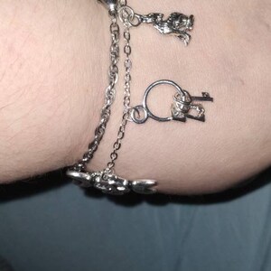 May include: A silver charm bracelet with three charms: a key, a heart, and a small animal figure. The bracelet is on a person's wrist.