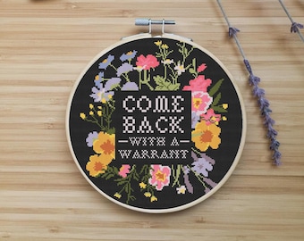 May include: A cross-stitch embroidery hoop with the text "COME BACK - WITH A - WARRANT" surrounded by colorful flowers on a black background. Three spools of thread and sprigs of lavender are arranged on a wooden surface.