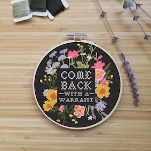 May include: A cross-stitch embroidery hoop with the text "COME BACK - WITH A - WARRANT" surrounded by colourful flowers on a black background. Three spools of thread and sprigs of lavender are arranged on a wooden surface.