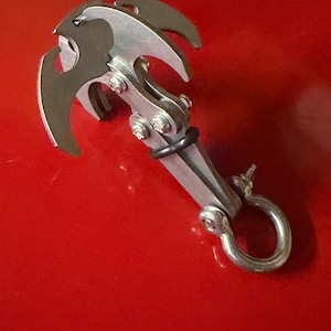 May include: A silver-colored grappling hook with three curved prongs and a ring at the base. The hook is made of metal and has a series of hinges and bolts. A black rubber ring is attached to the hook.