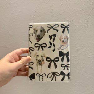 May include: Clear phone case featuring a pattern of black bows and images of dogs. The dogs are various breeds and sizes, some wearing bandanas. The case is held in a person's hand against a plain white background.