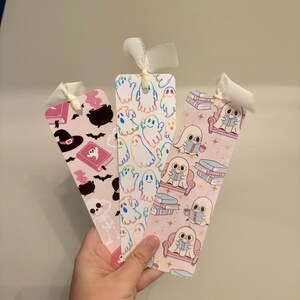 May include: Three decorative bookmarks with white ribbon ties. One bookmark features a witch hat, cauldron, and bats on a pink background. Another has ghost illustrations. The third shows ghosts reading books on a pink background.