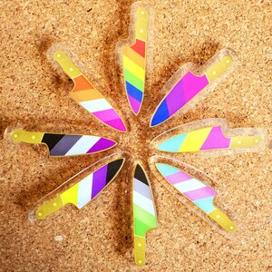 May include: A set of eight clear plastic knife-shaped ornaments, each featuring a different pride flag design. The flags include the rainbow, bisexual, transgender, non-binary, pansexual, lesbian, asexual, and genderfluid flags.