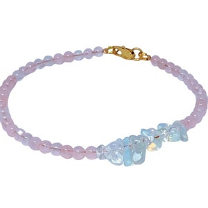 Pode incluir: Um delicado bracelete feito com contas cor-de-rosa e brancas. O bracelete tem um fecho dourado e um grupo de pedras brancas iridescentes.