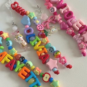 May include: Three colorful beaded phone charms featuring plastic letter beads spelling out names, including Olivia and Violet. The charms are decorated with various shapes such as stars, hearts, flowers, smiley faces, a cat in a glass, and a strawberry.