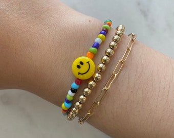 May include: Three bracelets: a beaded bracelet with a yellow smiley face, a gold beaded bracelet, and a gold chain link bracelet. The beaded bracelet features colorful beads. The gold bracelets have a shiny finish.