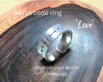 Puede incluir: Un anillo ajustable de plata con un mensaje personalizado en código Morse. El anillo está sobre una superficie de madera y el texto "Love" está escrito encima del anillo. El texto "custom your secret message" está escrito debajo del anillo.