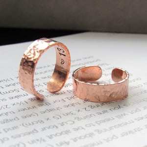 May include: Two hammered copper rings. One ring has the number "13" and a heart symbol engraved on the inside. The rings have a textured surface and an open design, allowing for adjustable sizing.