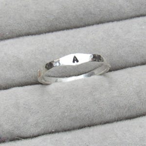 May include: A silver ring with a hammered texture and the letter "A" stamped on the top. The ring is displayed on a grey, textured surface, likely a jewellery display.