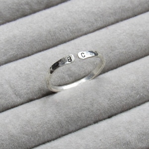 May include: A silver-colored ring with a hammered texture. The ring is open at the top and features the letters "B" and "C" stamped on the band. The ring is displayed on a gray, textured surface.