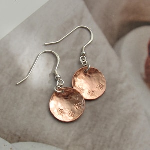 May include: A pair of copper-coloured disc earrings with silver-coloured ear wires. Each disc has a hammered texture and three small flower designs. The earrings are displayed on a neutral background.