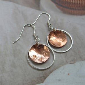 May include: Handmade earrings with copper discs, each adorned with floral patterns, set within silver hoops. The earrings have silver hooks and a warm, metallic colour scheme. These earrings are a unique accessory.