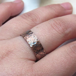 May include: A hammered copper ring with a textured surface. The ring is a band style, with a flat profile and a warm, reddish-brown colour. The hammered texture creates a unique, rustic appearance.