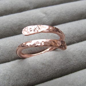 May include: A rose gold ring with a hammered finish. The ring has a spiral design that wraps around the finger. The ring is displayed on a grey, textured surface.
