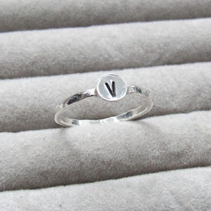 Op de afbeelding: Een zilveren ring met een gehamerde band en een cirkelvormige bedel. De bedel heeft de zwarte letter "V". De ring wordt gepresenteerd op een grijze, getextureerde ondergrond.