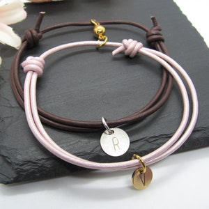 May include: Two leather bracelets. One is brown, the other is light pink. Each bracelet has a knotted closure. One bracelet has a silver charm with the letter "R", and the other has a gold charm.