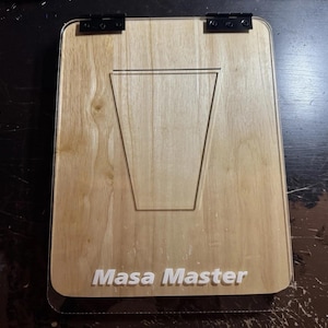 May include: A wooden clipboard with a light brown finish. The clipboard has a clear plastic overlay and black hinges. The words "Masa Master" are printed in white at the bottom. A cutout in the wood creates a stand.