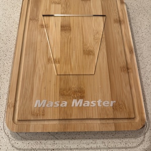 May include: A rectangular bamboo cutting board with a clear acrylic stand and a groove around the edge. The text "Masa Master" is printed in white. The board has black hinges.