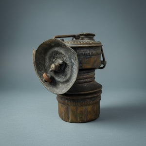 May include: An antique brass miner's lamp with a circular reflector and a handle. The lamp has a cylindrical body with a detailed design and a small, round base. The reflector is slightly angled, and the lamp shows signs of age and wear.