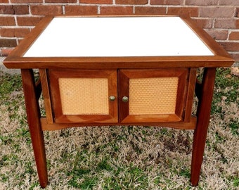May include: A mid-century modern end table with a white top and two doors with woven cane panels. The table is made of dark wood and has a simple, elegant design.