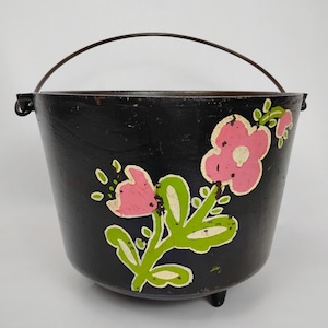 May include: A black cast iron pot with a curved handle and three small feet. The pot is decorated with pink flowers and green leaves. The pot is likely used for cooking.