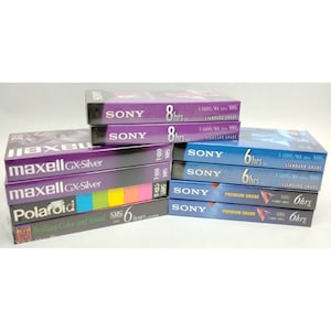 May include: A stack of VHS video tapes, including brands like Sony, Maxell, and Polaroid. The tapes are various colors, including blue, purple, and silver. The tapes are labeled with the brand name, recording time, and other information.