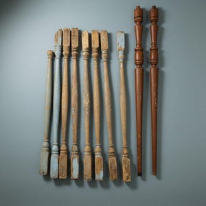 May include: A collection of ten wooden furniture legs arranged vertically. Eight legs feature a weathered, light-colored finish with traces of blue paint, while two legs on the right are finished in a solid, smooth reddish-brown wood tone.