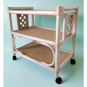 May include: A white and tan rattan serving cart with two shelves and a decorative lattice design on the sides. The cart has black wheels for easy mobility. The shelves are made of woven material.