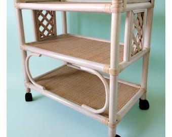 May include: A white and tan rattan serving cart with two shelves and a decorative lattice design on the sides. The cart has black wheels for easy mobility. The shelves are made of woven material.