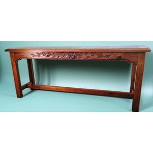 May include: A long, rectangular wooden table with a rich brown finish. The table features carved floral detailing along the top edge and sturdy, angled legs. A horizontal support beam connects the legs, adding to the table's stability. This piece is ideal for a living room or entryway.