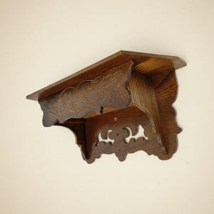 May include: A dark brown wooden wall shelf with carved floral details and decorative cutouts. The shelf has a layered design with a top shelf and a lower shelf, and is angled slightly to the left.