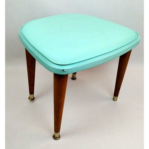 May include: A light blue cushioned stool with four wooden legs and gold metal tips. The stool has a square top and a rounded edge.