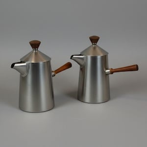 Può includere: Due caffettiere in metallo spazzolato con manici e coperchi in legno. Le caffettiere hanno una forma conica con un beccuccio per versare. I coperchi sono sormontati da un piccolo pomello in legno arrotondato. I manici sono in legno scuro.