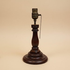 May include: A vintage table lamp with a dark brown wooden base and a brass lamp socket. The lamp has a classic design with a round base, a turned wooden column, and a pull chain for the light.