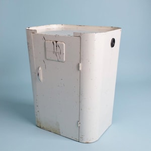 May include: A vintage, off-white metal cabinet with a curved design. The cabinet has a small door with a handle and a rectangular plate with text and dark streaks. A black circular hole is visible on the side. The cabinet is on a light blue background.