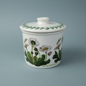 May include: White ceramic jar with lid, decorated with daisy illustrations and green foliage. The lid has a green leaf border. The jar is labeled "BELLIS PERENNIS" at the bottom. A charming piece for kitchen or home decor.