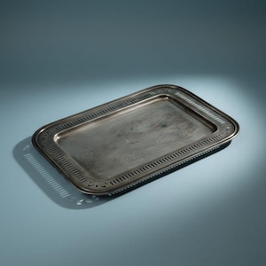 Puede incluir: Una bandeja rectangular de metal plateado que presenta una superficie central lisa y plana, junto con un borde decorativo con un patrón repetitivo de ranuras verticales y detalles sutiles de volutas en las cuatro esquinas redondeadas.
