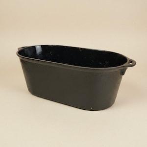 May include: A black, oval cast iron planter. The container has a slightly textured surface and small handles at each end. The interior is glossy black, and the exterior is matte black. The planter is designed to hold plants or other decorative items.