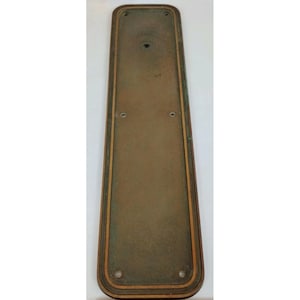 May include: A rectangular, antique brass door plate with rounded corners. The plate has a central panel with a slightly textured surface and a decorative border. There are four small holes and one larger hole visible on the plate.