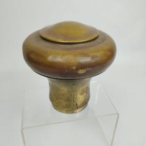 May include: A vintage brass door knob with a mushroom-shaped top and a cylindrical base. The knob has a tarnished, aged appearance, with variations in color from gold to brown. The base has a textured surface.