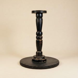 May include: A black, wooden pedestal stand with a round base and a smaller, circular top. The stand's central column features a turned design with a series of rounded sections. The wood shows signs of wear, adding to its vintage aesthetic. Ideal for displaying decorative items.