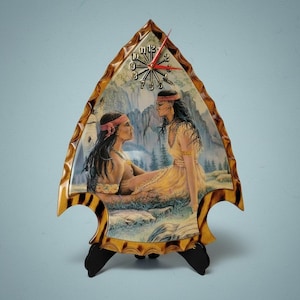 May include: Arrowhead-shaped clock with a painting of two women in Native American attire. The clock face has black numbers and red hands. The frame has a brown and yellow tortoiseshell pattern, and the clock stands on a black base.