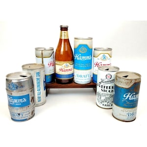 Könnte beinhalten: Eine Sammlung von Vintage-Bierdosen und -flaschen von Hamm's. Die Dosen sind in verschiedenen Farben und zeigen das Hamm's-Logo und den Slogan. Die Flaschen sind aus klarem Glas und haben ein grünes Etikett mit dem Hamm's-Logo.