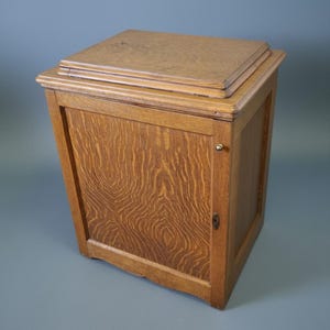 May include: A wooden cabinet with a rectangular shape and a hinged door. The cabinet is made of light brown wood with a natural wood grain pattern. The top of the cabinet has a layered design.