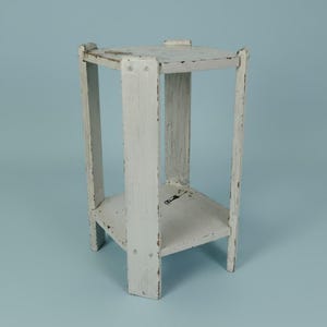 May include: A white wooden plant stand featuring a hexagonal top and a matching lower shelf. The structure is supported by three vertical legs, with visible distressed paint finish showing wear and dark wood grain underneath the white surface.