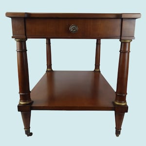 Puede incluir: Una mesa auxiliar de madera marrón oscuro con un cajón y un estante inferior. La mesa tiene cuatro patas decorativas con detalles dorados y pequeñas ruedas. El cajón presenta un pomo redondo de metal ornamentado. La mesa mide aproximadamente 61 cm de ancho, 46 cm de profundidad y 71 cm de alto.