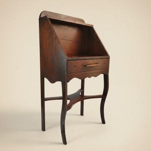 May include: A dark brown wooden writing desk featuring a slanted top surface, a single horizontal drawer with a wooden handle, and four curved legs connected by a central X-shaped support brace. The desk has a scalloped apron below the drawer.