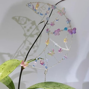 May include: A crescent moon-shaped plant trellis crafted from silver wire, adorned with iridescent butterfly-shaped beads and small colorful star-shaped beads. The structure features a delicate, open-work design with beads strung along the wire frame.
