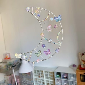 以下が含まれることがあります： 銀色の金属フレームで作られた三日月形のワイヤーオーナメント。パステルカラーの半透明なビーズで装飾されており、月の中に蝶や星の形をした小さなチャームが吊り下げられているデザインです。